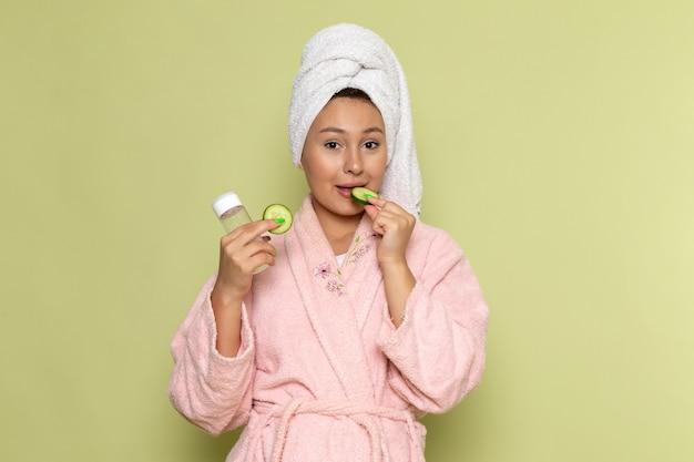 Woman in pink robe eating cucumber slice and holding skincare products