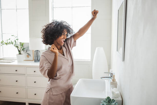 Alt text: Happy African American woman dancing in a modern bathroom, celebrating in front of a sink and mirror.